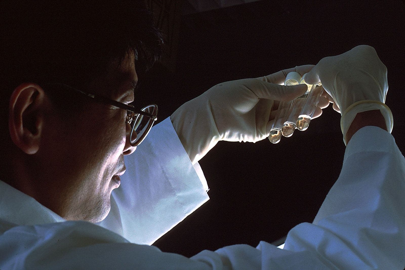 A researcher in gloves holds a small vial up to the light